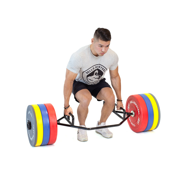 A person in a gray T-shirt and black shorts readies to lift the Bells of Steel Open Trap Bar adorned with colorful plates. Their determined stance contrasts the minimal white setting, highlighting the vibrant equipment.