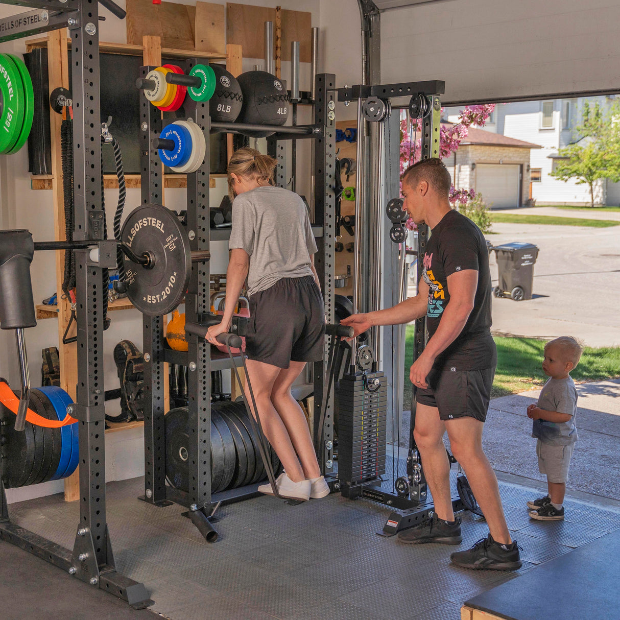 A woman works out on a dip machine in a garage gym featuring the Hydra Storage Builder by Bells of Steel. A supportive man stands nearby as a child watches. The gym is equipped with organized weightlifting gear, and beyond, the sunny street is visible.