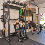 A woman does sit-ups on the Home Gym Builder by Bells of Steel USA, smiling at a man who encourages her in their garage gym, surrounded by power racks and weight plate sets.