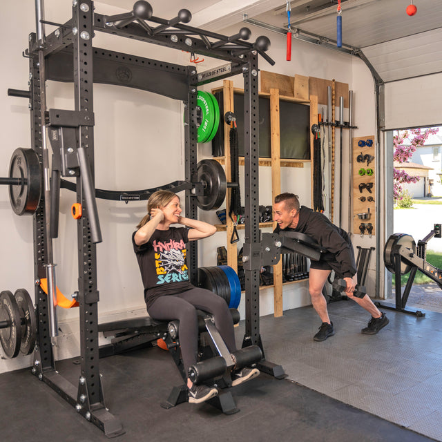 A woman does sit-ups on the Home Gym Builder by Bells of Steel USA, smiling at a man who encourages her in their garage gym, surrounded by power racks and weight plate sets.