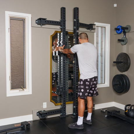 A person adjusts the Bells of Steel Manticore Folding Half Rack Builder (3" x 3", 1" holes) in a home gym with a large mirror, weight plates, tan walls, black flooring, two windows, and other equipment.