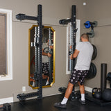 A man in a gray t-shirt and patterned shorts adjusts the Bells of Steel Manticore Folding Half Rack Builder (3" x 3", 1" Holes) in a home gym with a mirror, weight plates, barbells, kettlebells, and other fitness equipment.