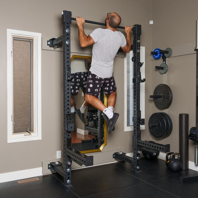 A man in a gray shirt and patterned shorts does a pull-up on the Bells of Steel Manticore Folding Half Rack Builder (3" x 3", 1" Holes) in his home gym, surrounded by weight plates, a kettlebell, and other fitness gear.