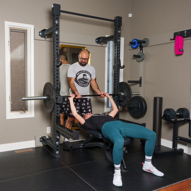 Wearing teal leggings and white sneakers, a person bench presses on the Bells of Steel Manticore Folding Half Rack Builder (3" x 3", 1" Holes) as a trainer in shorts and a t-shirt stands behind to spot and provide support.