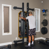 A person adjusts a Bells of Steel Manticore Folding Half Rack (3" x 3", 1" Holes) in a bright room. Weight plates are stored on a nearby rack, with two tall windows letting in natural light.
