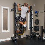 A man in a gray t-shirt and patterned shorts performs a pull-up on the Bells of Steel Manticore Folding Half Rack (3"x3", 1" holes) in a home gym, surrounded by free weights, plates, and mirrors.