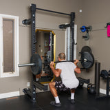 A man in workout clothes squats with a loaded barbell in a home gym, using the Bells of Steel Manticore Folding Half Rack (3" x 3", 1" holes); various weights and a kettlebell appear in the background.