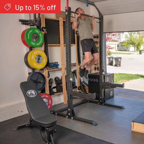 A man does pull-ups on the Bells of Steel USA Home Gym Builder in a garage gym with weights, bench, and equipment; a red “Up to 15% off” sale banner is displayed in the top left corner.