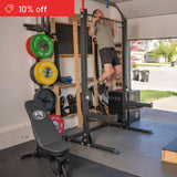 A man uses the Bells of Steel USA Home Gym Builder power rack for a pull-up in a home garage gym with barbell, bench, and weight plates. A “10% off” banner appears in the top left corner of the image.