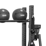 Two black 10-pound medicine balls sit on a metal rack, while a battle rope hangs from a Bells of Steel Utility Hook attached to the exercise equipment frame, highlighting organized gym storage.