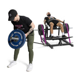 In a plain white room, one person in black clothing lifts a barbell with blue weights while another uses the Bells of Steel Hip Thrust Machine.