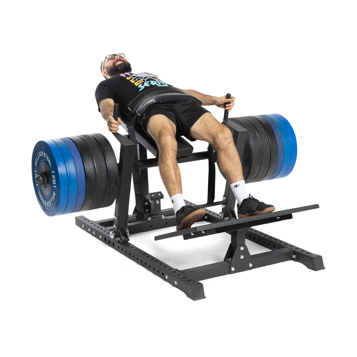 A person is using a weight-loaded leg press machine in a black T-shirt, shorts, and socks with black shoes. The machine, next to a Bells of Steel Hip Thrust Machine, has blue and black plates. They look relaxed and are smiling.