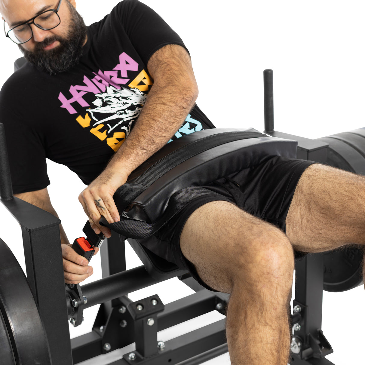 Seated at a Bells of Steel Hip Thrust Machine, a bearded person in glasses, a black graphic tee, and shorts secures their belt as resistance bands hang nearby against the white backdrop.