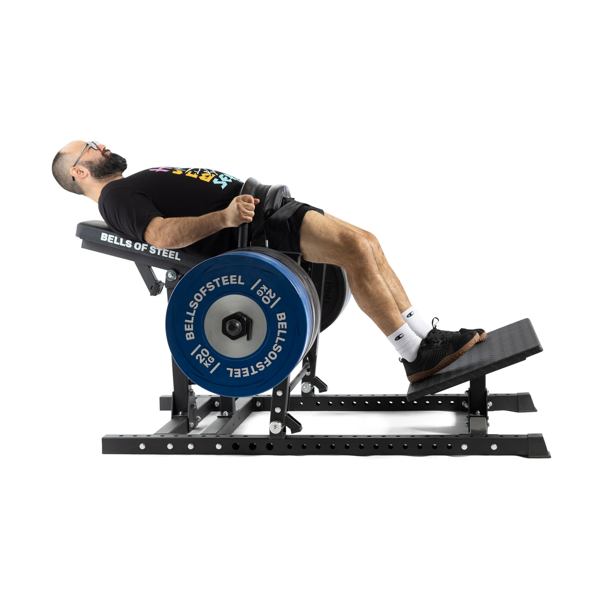 A bearded man in glasses uses a Hip Thrust Machine. Wearing a black t-shirt, shorts, and sneakers, he adds resistance bands to his workout. The machine includes blue weights marked with "Bells of Steel.