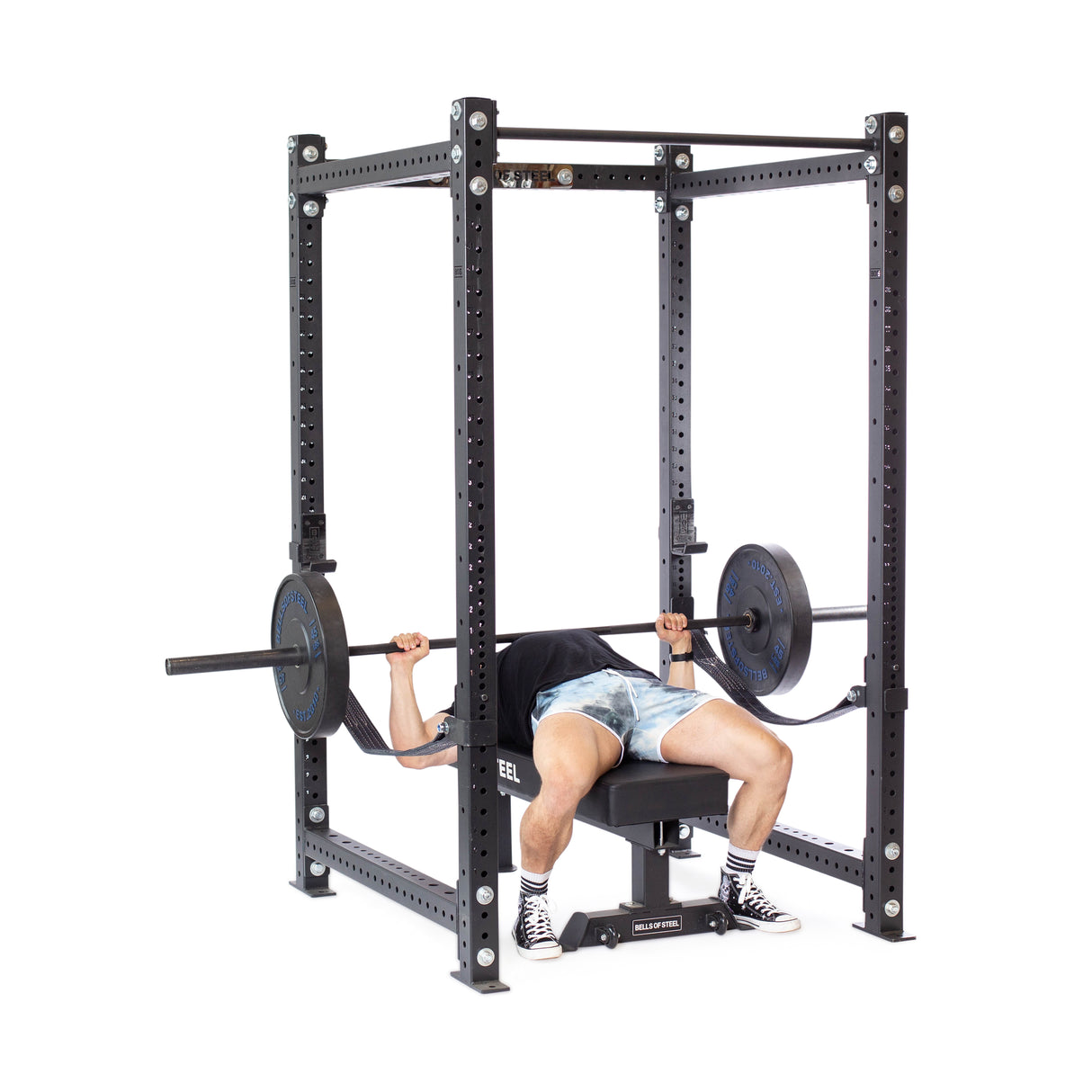 A person bench presses a barbell in the Bells of Steel Hydra Four Post Power Rack Builder. They're wearing a black shirt, blue tie-dye shorts, and striped sneakers. The sleek black rack, with modular safety components, enhances this dynamic home gym setup.