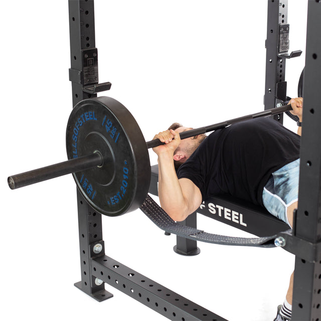 A person in a black shirt is lying on a bench within the Bells of Steel Hydra Four Post Power Rack Builder (3" x 3", ⅝" Holes), performing a bench press with large black weight plates, surrounded by modular components typical of an advanced home gym.