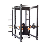 A person in black workout attire and a cap performs a squat with a barbell loaded with weights inside the Bells of Steel Hydra Six Post Power Rack Builder (3" x 3", ⅝" Holes). The sturdy frame features modular components, multiple adjustment holes, and Hydra rack attachments.