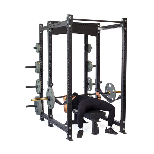 Inside the Bells of Steel Hydra Six Post Power Rack Builder, a person in black workout gear confidently bench presses a weighted barbell. The rack, featuring modular components and weight plates on its sides, supports their intense workout.