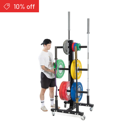 Wearing athletic gear, a person loads a green plate onto the Bells of Steel Hydra Bumper Plate Tree Builder, which holds colorful plates and two barbells. A red "10% off" label appears in the top left corner.