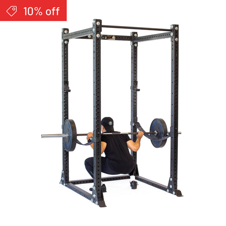 Wearing black gym clothes and a hat, someone squats with a barbell inside the Bells of Steel Hydra Flat Foot Power Rack Builder (3" x 3", ⅝" Holes). A red "10% off" sale banner appears—ideal for upgrading your home gym!.