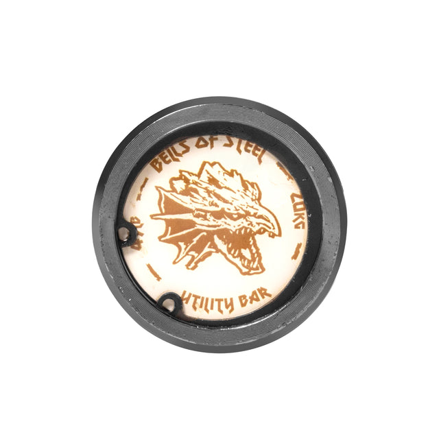 A close-up of a round metal plate with an engraved dragon head design displays "Bells of Steel" and "Multi-Purpose Olympic Barbell – The Utility Bar," set in a sleek black circular frame, ideal for powerlifting enthusiasts.