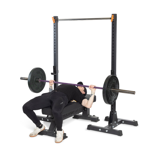 A powerlifter wearing black athletic clothing and white sneakers is on a bench press, lifting a Bells of Steel Multi-Purpose Olympic Barbell – The Utility Bar loaded with weights. The bench rests on an adjustable rack as the athlete focuses intently on their routine.