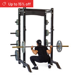 A person in black workout clothes squats with a barbell inside the Bells of Steel Hydra Collegiate Power Rack Builder (3" x 3", ⅝" Holes), loaded with weights. A red banner in the corner says "Up to 15% off." Ideal for any home gym.