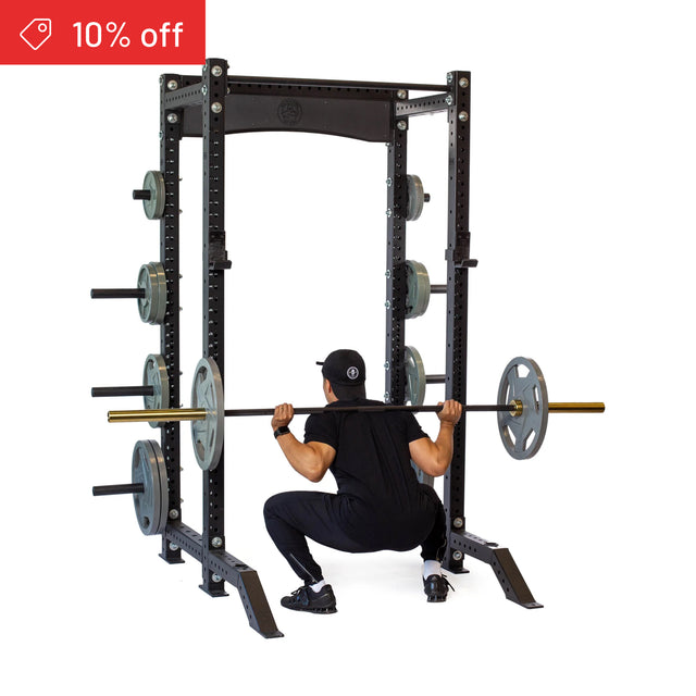 A person in black workout gear and a cap does a squat with a barbell in the Bells of Steel Hydra Collegiate Power Rack Builder (3" x 3", ⅝" Holes), with plates stored on the rack and a "10% off" sale tag in the top left corner.