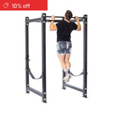 A person in a black shirt, tie-dye shorts, and striped socks does a pull-up on a Bells of Steel Hydra Four Post Power Rack Builder (3"x3", ⅝" holes) in a white home gym. A red "10% off" banner appears in the top left corner.