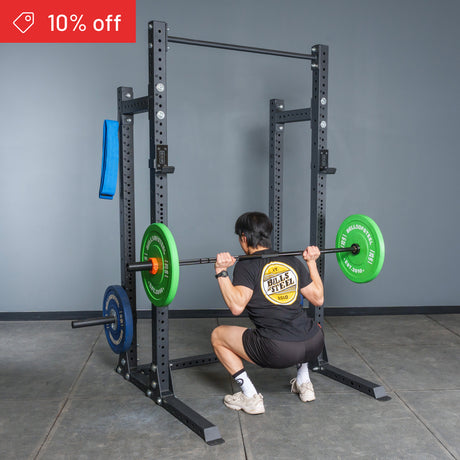 A person does a barbell squat inside the Bells of Steel Hydra Half Rack Builder (3" x 3", ⅝" Holes) with weight plates. A red "10% off" banner appears upper left. The gym has gray walls and a concrete floor. Perfect for home gyms!.