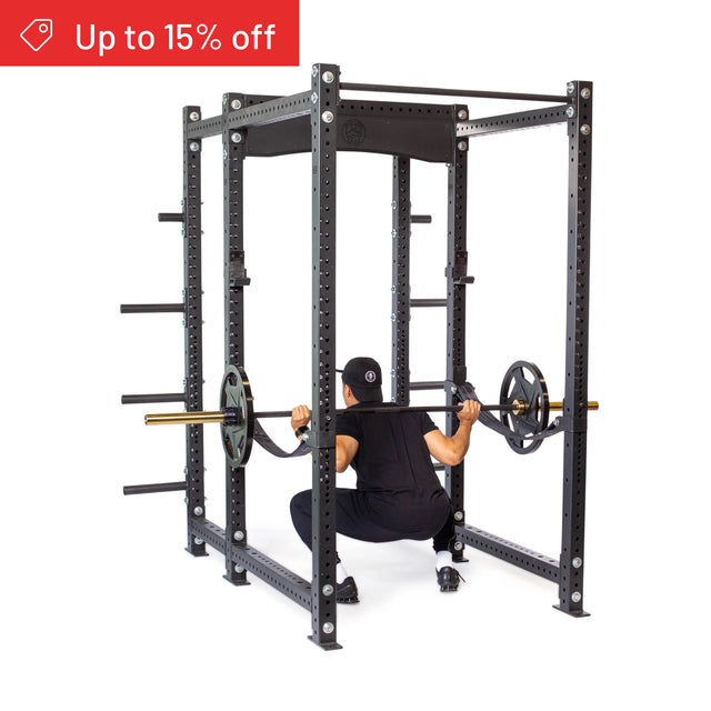 A person squats with a barbell inside a black Bells of Steel Hydra Six Post Power Rack Builder (3" x 3", ⅝" Holes) featuring weight plate storage arms and adjustable safety pins. A red banner in the top left says "Up to 15% off.