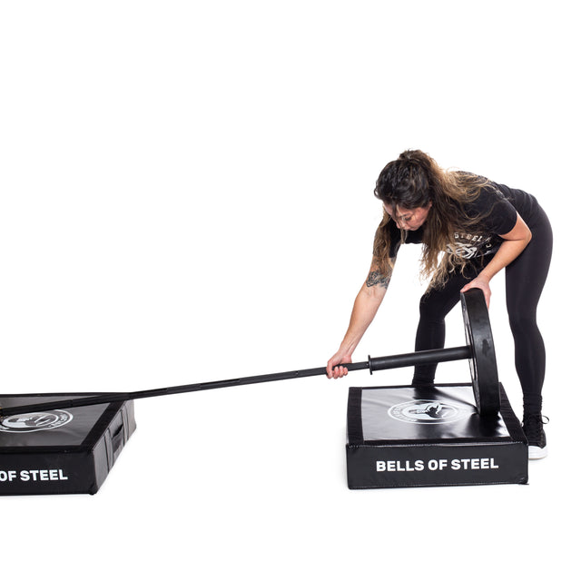 A woman in a black workout outfit adjusts weights on a barbell resting on Bells of Steel Deadlift Pads, bending forward and focused.