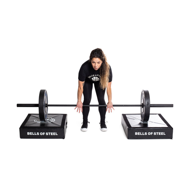 A woman in athletic attire stands on padded platforms labeled "Bells of Steel" while lifting a barbell. Surrounded by Deadlift Pads (Pair) for silencing, she remains focused against a plain white background.