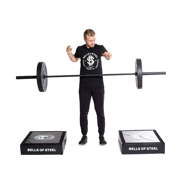 A man demonstrates a lift between two black Bells of Steel platforms on a white backdrop, using a barbell and Deadlift Pads to reduce noise and protect the floor. He's dressed in a black t-shirt and pants.