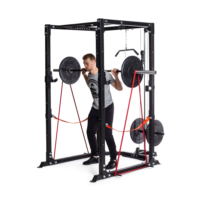 A person powerlifting in a black squat rack holds a barbell with weights on their shoulders. Bells of Steel's Fabric Non-Slip Resistance Bands (41") add resistance by connecting the barbell to the base.