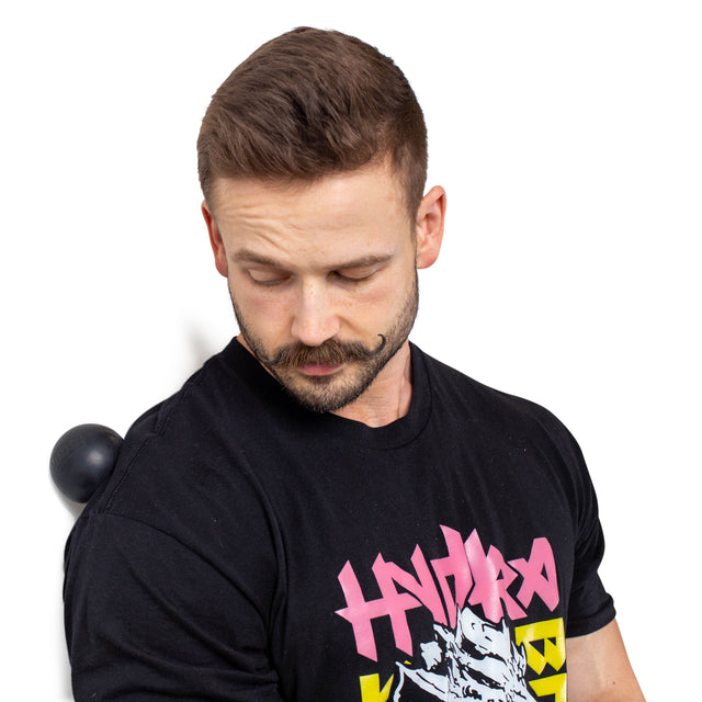 A man with a short beard and styled hair uses Bells of Steel Massage Balls, an essential mobility tool, against a wall for back pressure. Wearing a black graphic T-shirt, he maintains focus during this effective muscle massage.