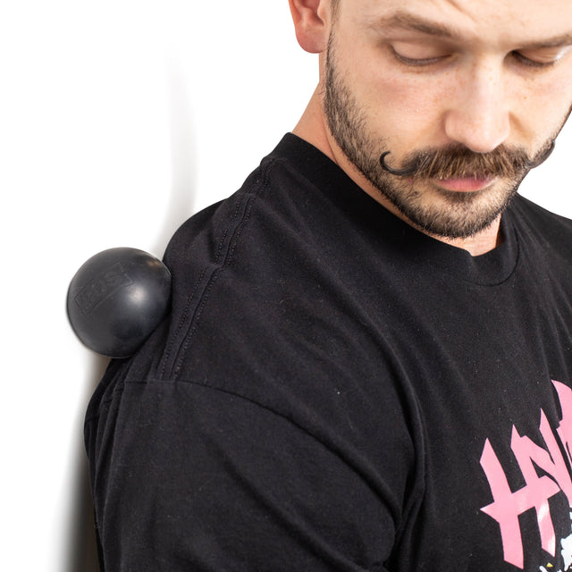 A man with a mustache uses Bells of Steel's black Massage Balls, an ideal mobility tool, against a white wall to ease shoulder tension. He wears a black T-shirt with pink lettering.