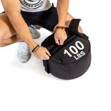 A person in athletic wear crouches and grips the handle of a Bells of Steel Fitness Sandbag Set labeled "100 LBS"—perfect for sandbag workouts—against a white background.