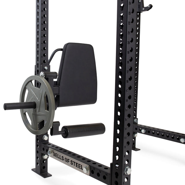 Close-up of a black squat rack featuring a pulley system, resistance bands, and a "Bells of Steel" footplate. It has multiple holes for adjustable heights and can include the Bells of Steel Leg Curl / Leg Extension Rack Attachment with a handle and weight-loaded pulley.