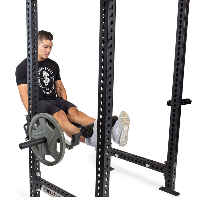 A man in a black T-shirt and shorts uses a Bells of Steel Leg Curl/Leg Extension Rack Attachment in the gym, which boasts an impressive weight plate capacity, staying focused on his workout with plates attached.