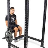 A man uses the Bells of Steel Leg Curl / Leg Extension Rack Attachment, seated with a weight plate loaded near his knees—an ideal choice for any home gym setup.