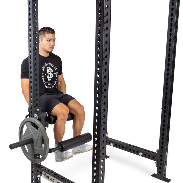 A person sits on a bench inside a Bells of Steel power rack, gripping the seat and preparing to use the Leg Curl / Leg Extension Rack Attachment with weight plates, perfect for any home gym. They are wearing a black T-shirt and shorts.