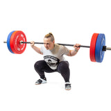 A person in a gray "Bells of Steel" T-shirt, black leggings, and gym shoes performs a squat with a barbell loaded with Colored Bumper Plates featuring stainless steel inserts. The gym backdrop is pristine white.