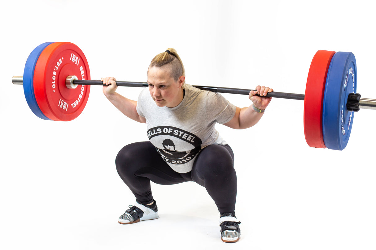 Wearing a "Bells of Steel" shirt, an individual performs a barbell squat with the Multi-Purpose Olympic Barbell – The Utility Bar, using red and blue plates. Against a plain white background, their focused expression and athletic stance highlight dedication to powerlifting precision.