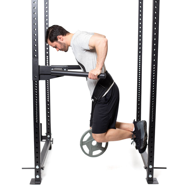 A man performs weighted dips on a dip bar in a gray t-shirt and black shorts, using the Bells of Steel Belt Squat Belt with a large plate hanging from his waist, all set against a plain white background.