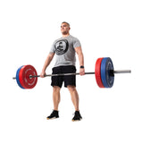 A man in a gray t-shirt and black shorts holds a barbell with Bells of Steel Colored Bumper Plates, featuring durable stainless steel inserts. Standing on a white background, he wears black shoes and has a focused expression.