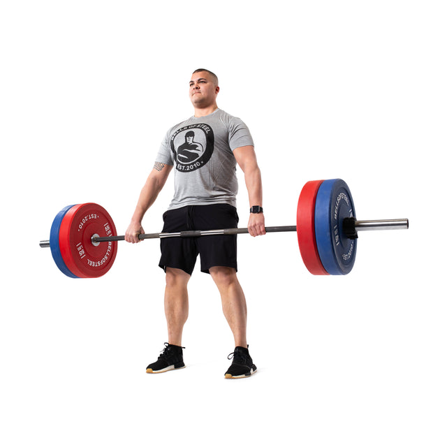 A man in a gray t-shirt and black shorts deadlifts a barbell loaded with Bells of Steel Colored Bumper Plate Sets, featuring stainless steel inserts, against a white background.