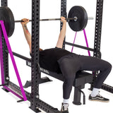 Wearing black workout clothes, a person bench presses on a weight bench with a barbell inside a power rack equipped with purple safety straps and Bells of Steel Standard Band Pegs for added support.