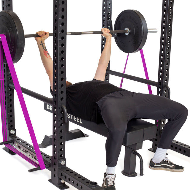 Wearing black workout clothes, a person bench presses on a weight bench with a barbell inside a power rack equipped with purple safety straps and Bells of Steel Standard Band Pegs for added support.