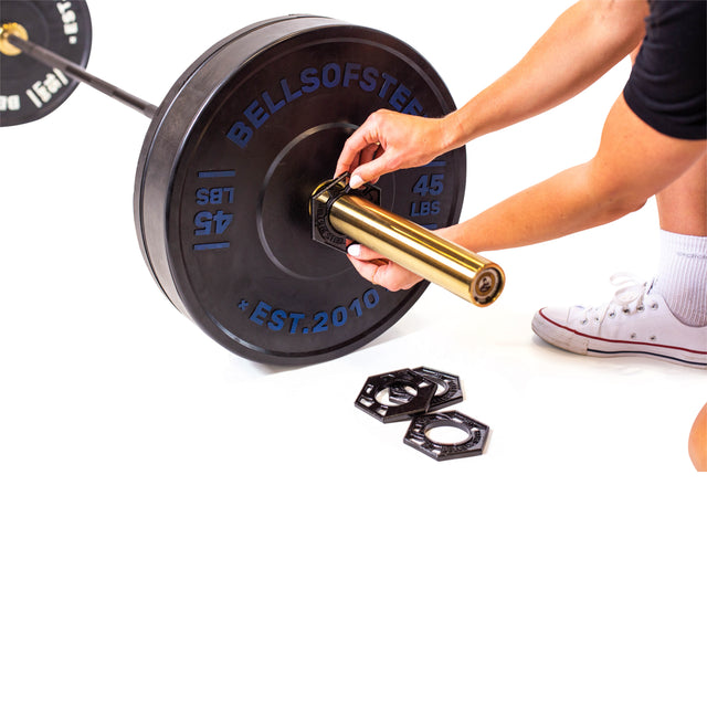 A person kneels beside an Olympic barbell with 45-pound weights, adjusting a collar. Nearby are two hexagonal collars and Bells of Steel Fractional Iron Plates - 0.5 LB. They wear white sneakers.